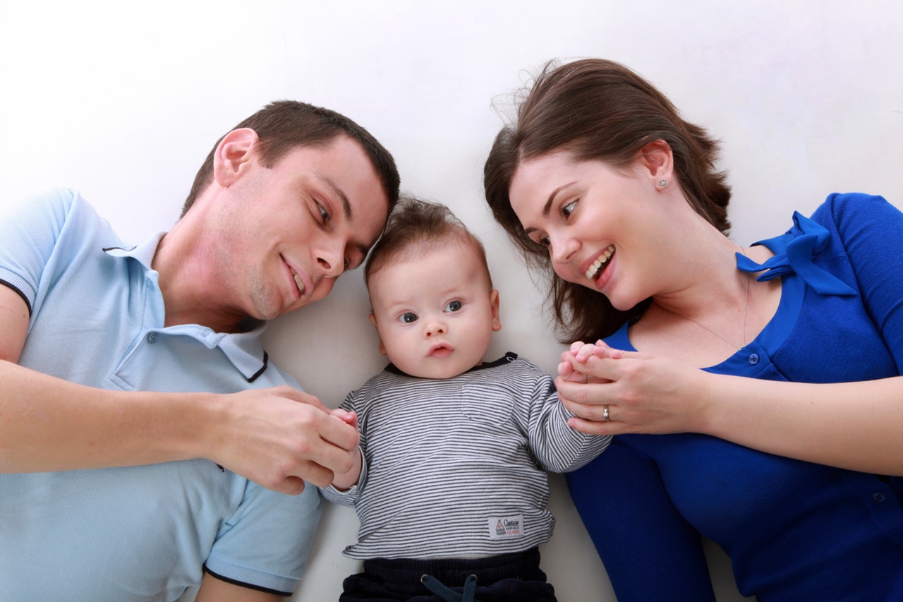 Zdjęcie przedstawia uśmiechniętą rodzinę. Leży kobieta, dziecko i mężczyzna.. Dorośli trzymają dziecko za ręce.